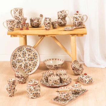 Service de vaisselle en grès artisanal peint à la main avec des motifs floraux colorés, incluant des gobelets, une théière, des bols et une grande assiette.