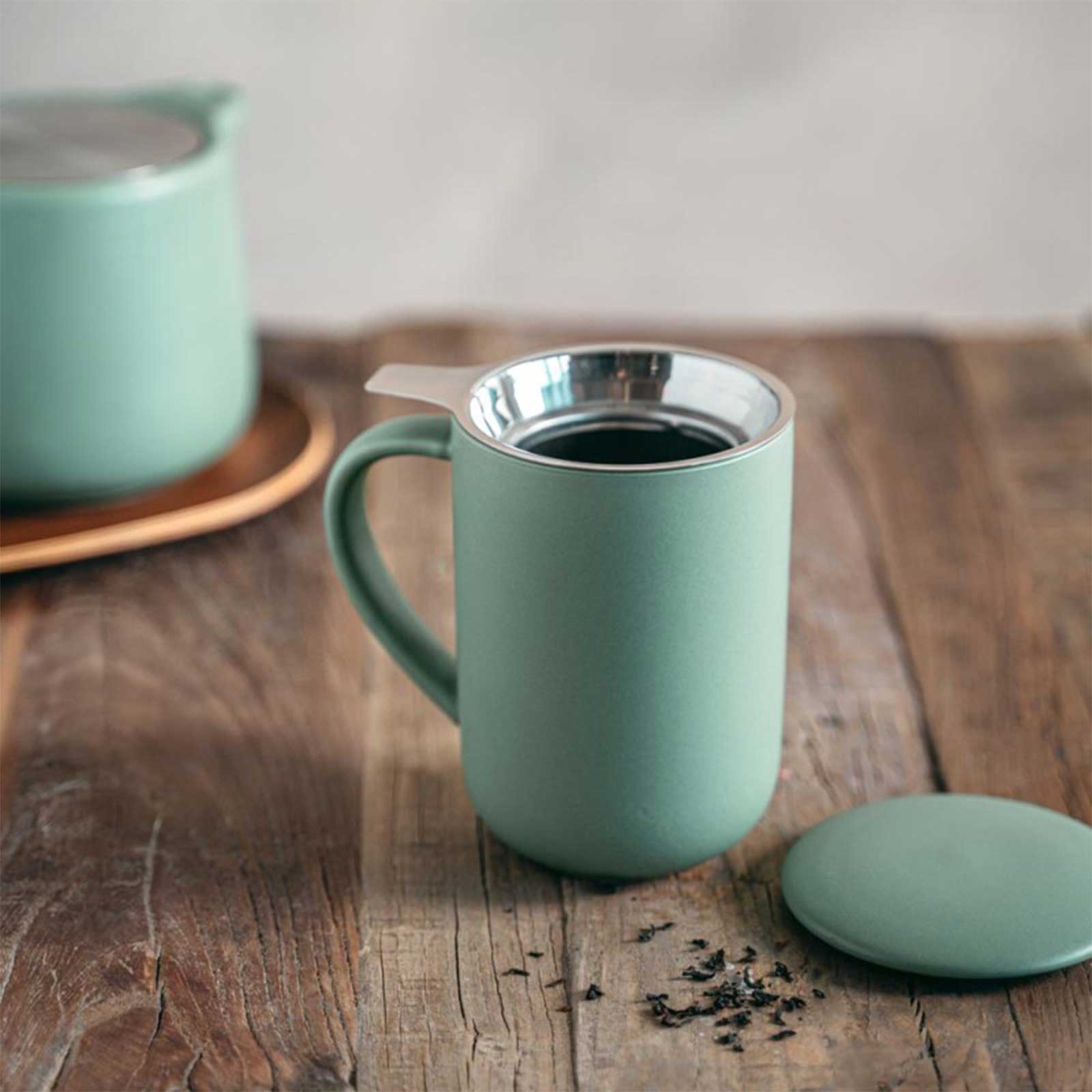 Tisanière en grès amande mat avec infuseur en métal argenté, posée sur une table en bois avec quelques feuilles de thé éparpillées