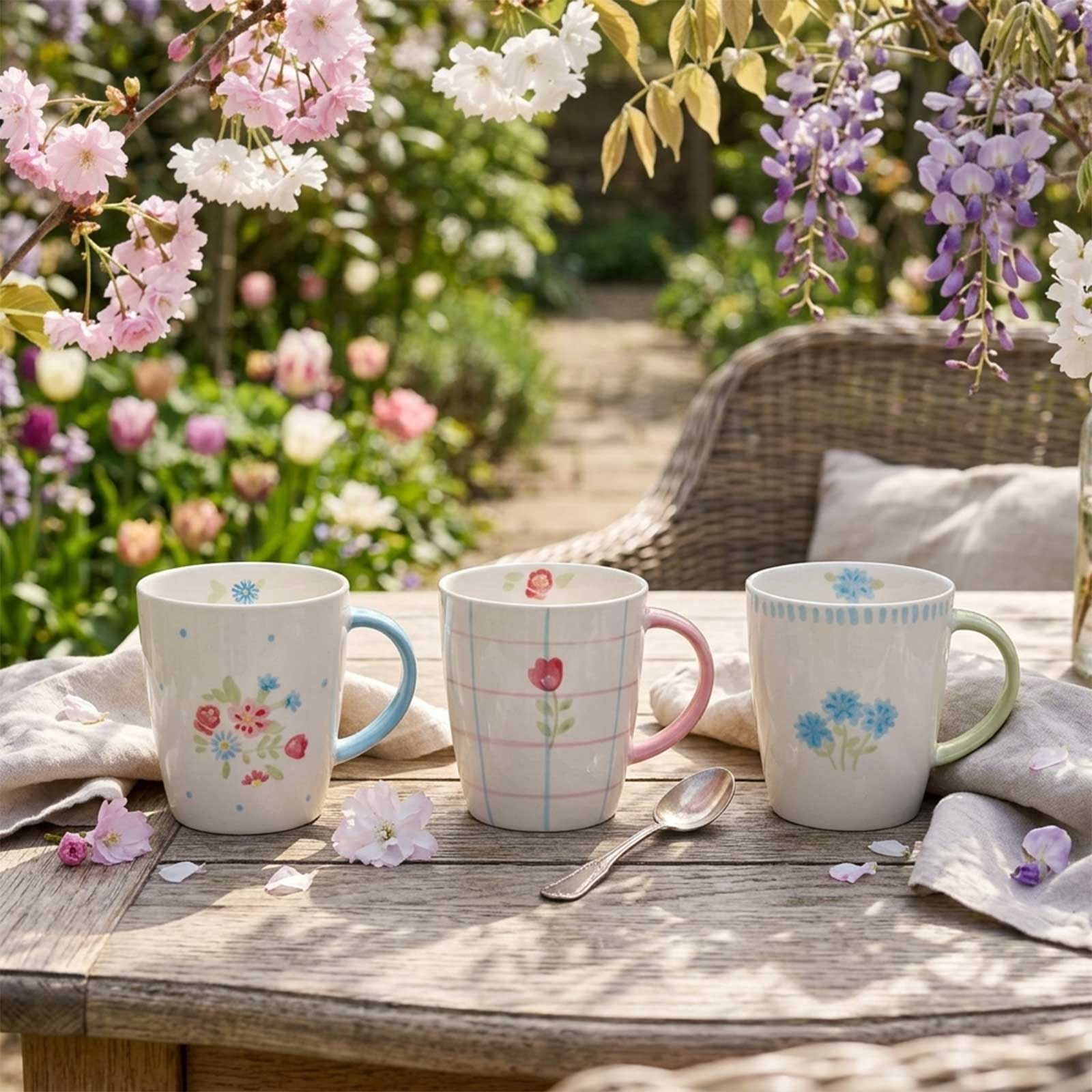 Trois mugs en grès émaillé artisanal avec motifs floraux délicats et anses colorées, disposés sur une table en bois dans un jardin fleuri