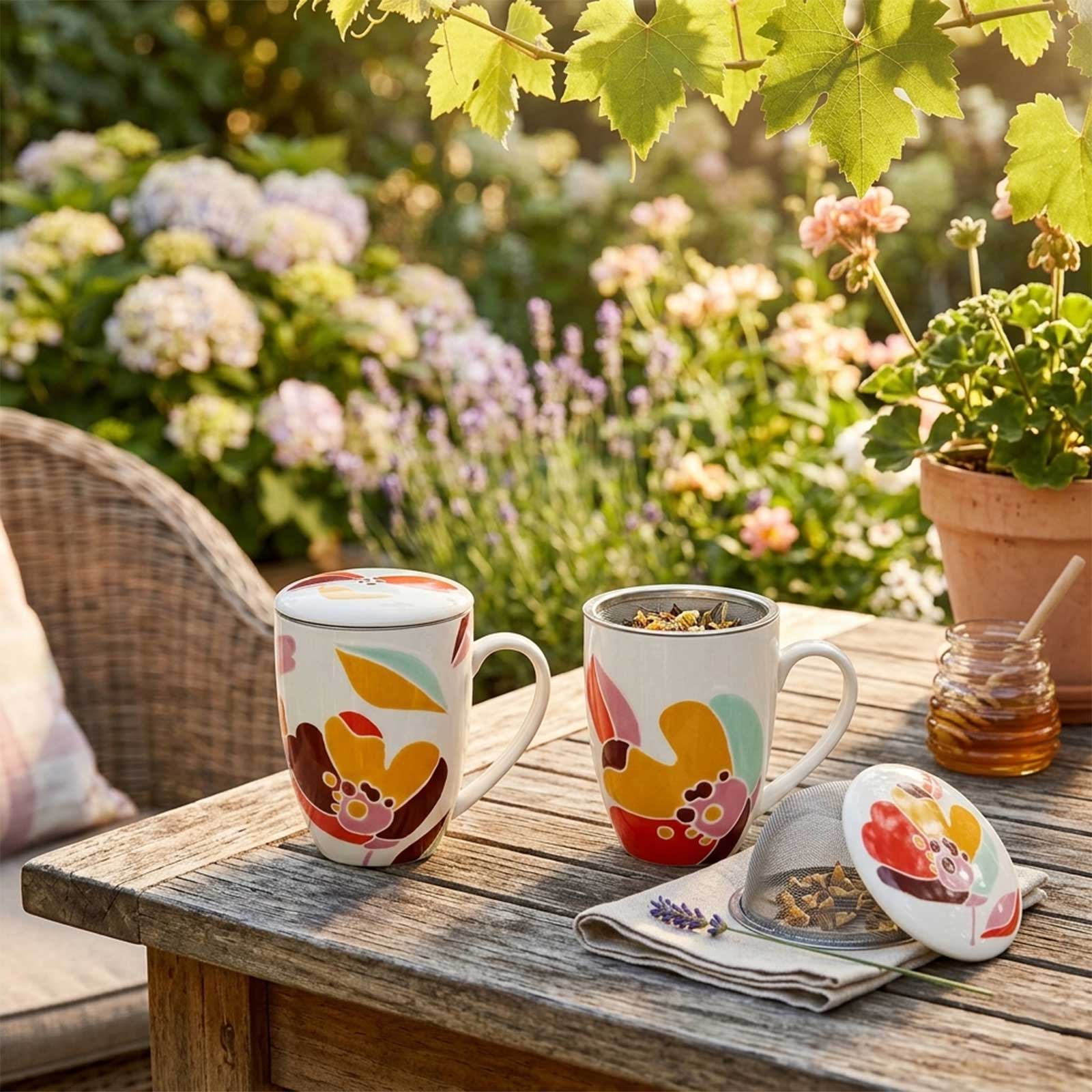 Deux tasses à tisane Amadeus Alexia avec motif floral coloré, disposées sur une table en bois dans un jardin fleuri, avec un petit pot de miel à côté