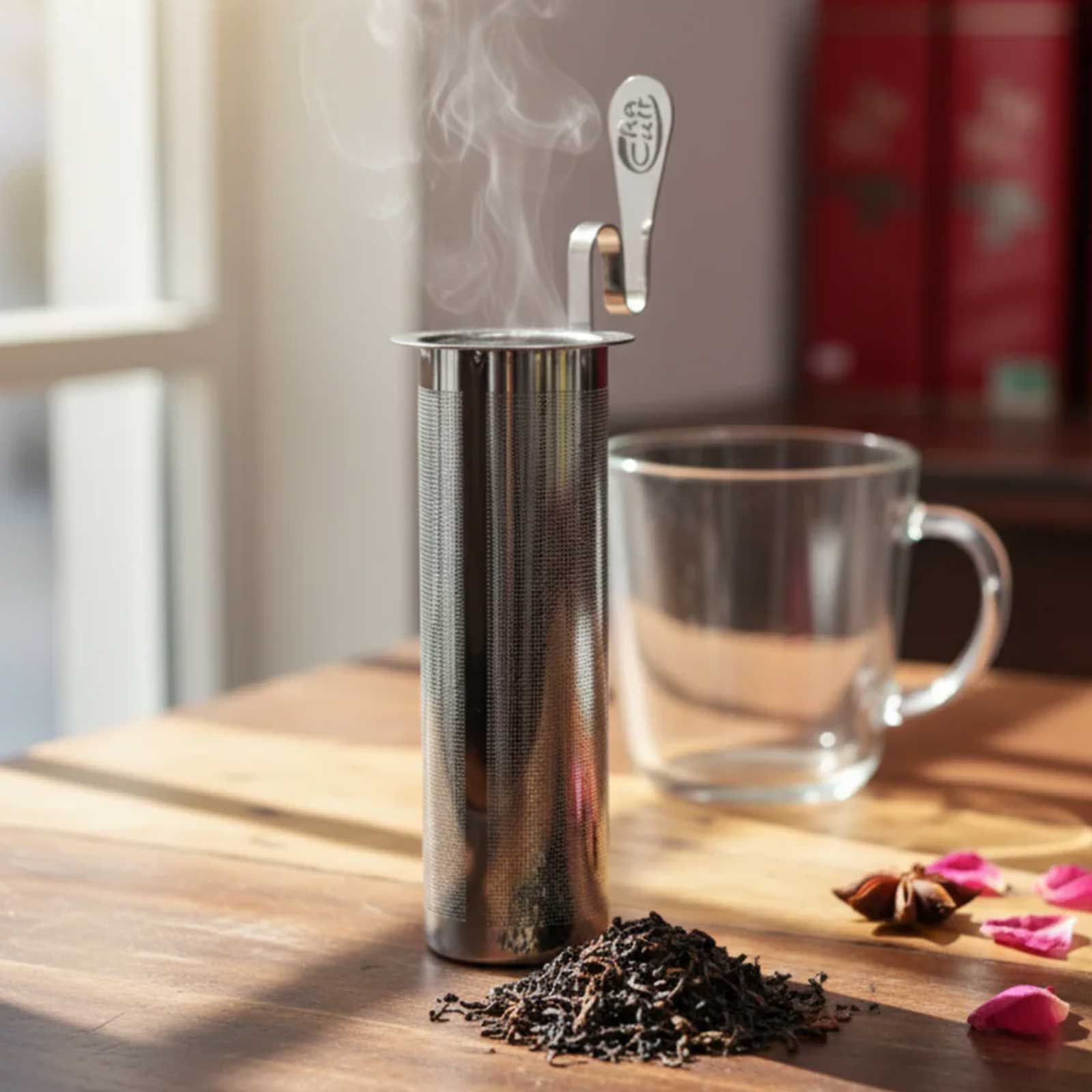 Filtre à thé cylindrique en acier inoxydable avec une cuillère gravée 'Cha Cult' sortant de la vapeur, à côté d'une tasse en verre vide et d'une petite pile de thé noir sur une table en bois.