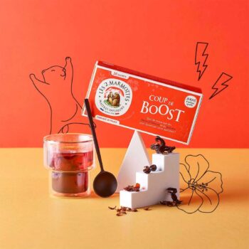 Boîte de sachets d'infusion Les 2 Marmottes Coup de Boost rouge, posée sur un support blanc, à côté d'un verre de boisson rouge et d'une cuillère.