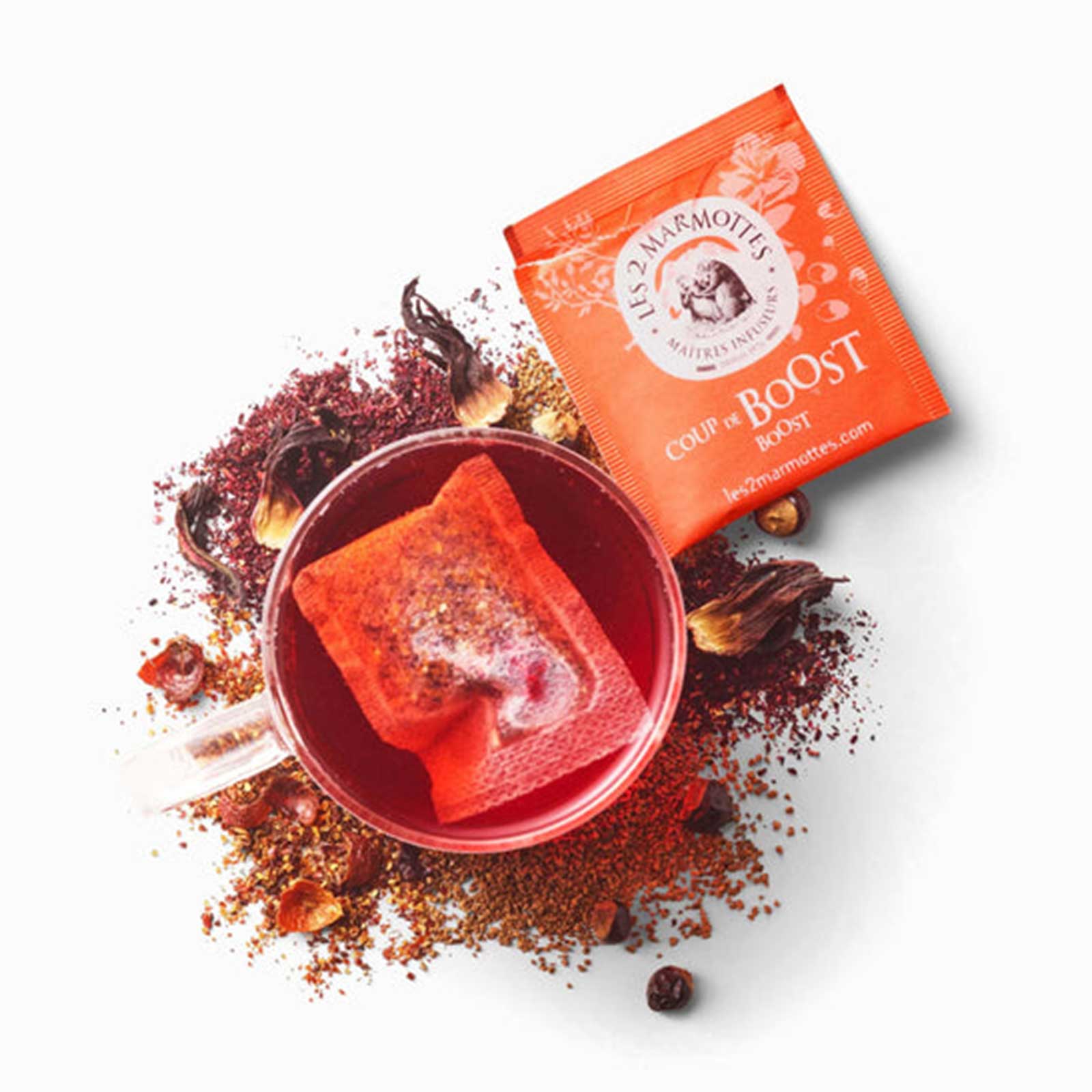 Sachet d'infusion Les 2 Marmottes Coup de Boost rouge orangé dans une tasse remplie d'infusion, entouré de plantes sèches