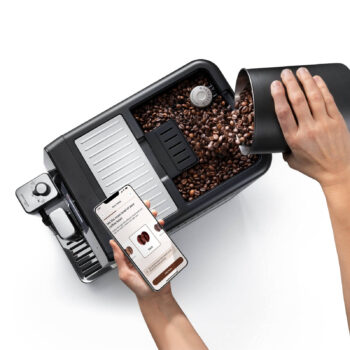 Main versant des grains de café dans le réservoir d'une machine à espresso De'Longhi Eletta Ultra, avec un smartphone affichant l'application Bean Adapt