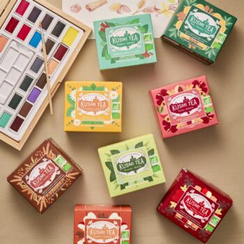 Plusieurs boîtes de thés et infusions Kusmi Tea disposées autour d'une palette d'aquarelle, dont une boîte pêche abricot au centre
