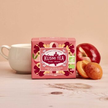 Boîte de Kusmi Tea infusion pêche-abricot sans théine, avec une tasse blanche et des fruits frais à côté