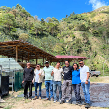 Groupe de personnes souriantes posant devant une plantation de café au Pérou, avec des structures de séchage et des collines verdoyantes en arrière-plan