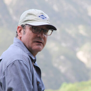Portrait d'Arnaud Causse portant une casquette beige avec le logo "BERG" et des lunettes, sur fond de montagnes floues.