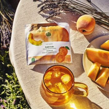 Sachet de thé glacé Palais des Thés Plein Sud avec des fruits (abricot, melon) et un verre de thé glacé rempli de glaçons, sur une nappe en lin.