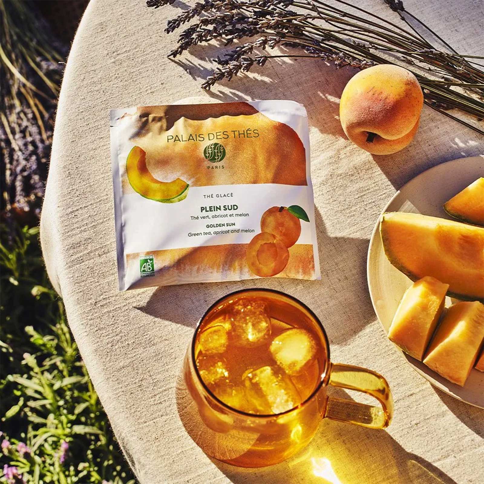 Sachet de thé glacé Palais des Thés Plein Sud avec des fruits (abricot, melon) et un verre de thé glacé rempli de glaçons, sur une nappe en lin.