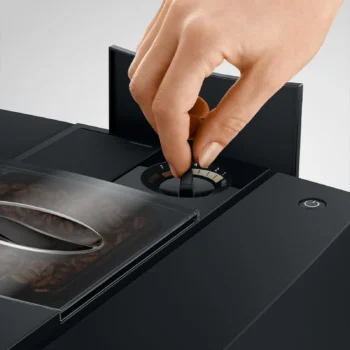 Main ajustant le bouton de réglage de l'intensité du café sur une machine JURA E8, avec des grains de café visibles dans le réservoir