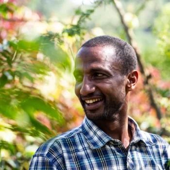 Khalid Shifa, producteur de café éthiopien, souriant dans un environnement verdoyant, portant une chemise à carreaux bleus et blancs