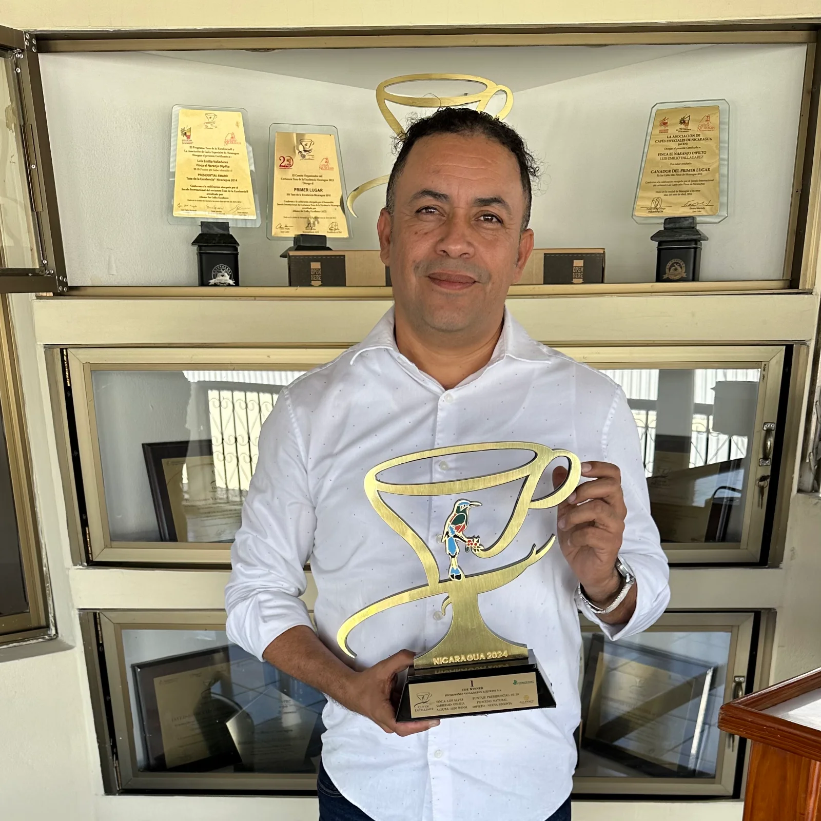 Producteur de café posant fièrement avec le trophée Cup of Excellence Nicaragua 2024 devant une vitrine remplie de distinctions
