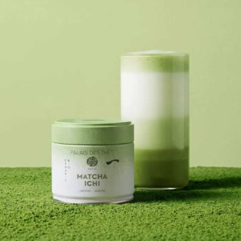 Boîte de thé Matcha Ichi de Palais des Thés posée devant un verre rempli d'un latte au matcha aux nuances de vert et de blanc sur fond végétal