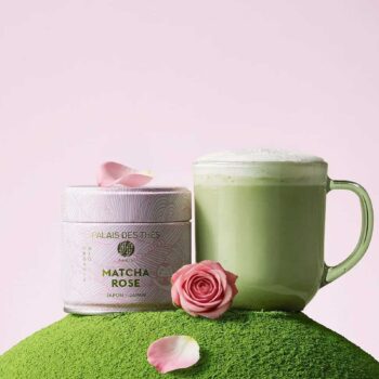 Boîte métallique de Matcha Rose Palais des Thes posée sur un monticule de poudre de thé vert, accompagnée d'un mug de matcha latte et de pétales de rose