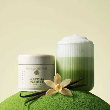Boîte de Matcha Vanille Palais des Thés posée sur un monticule de thé en poudre vert, à côté d'un verre cannelé de matcha latte mousseux et vanille