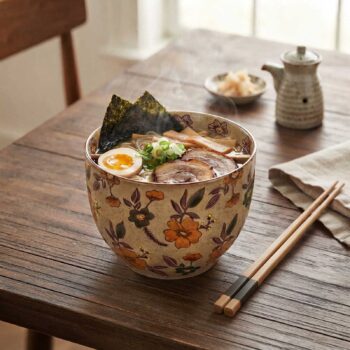 Bol à ramen en céramique orné de motifs floraux colorés, rempli de nouilles, œuf mollet, porc chashu et nori, posé sur une table en bois avec baguettes