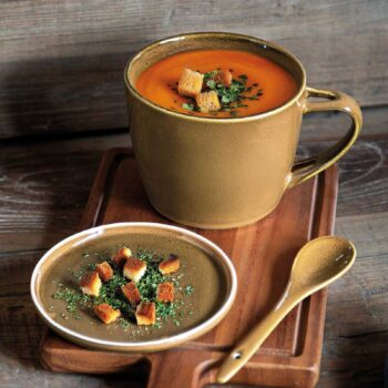 Grande tasse en céramique ocre remplie de soupe de tomate et de croûtons sur un plateau en bois avec une petite assiette assortie et une cuillère