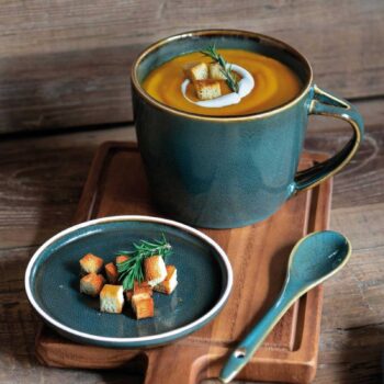 Grande tasse en grès bleu vert remplie de soupe onctueuse garnie de crème fraîche, d'herbes aromatiques et de croûtons sur une planche en bois