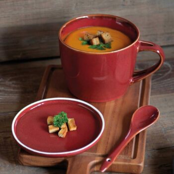 Mug en céramique rouge avec soupe veloutée et croûtons, accompagné d'une petite assiette assortie et d'une cuillère sur un plateau en bois