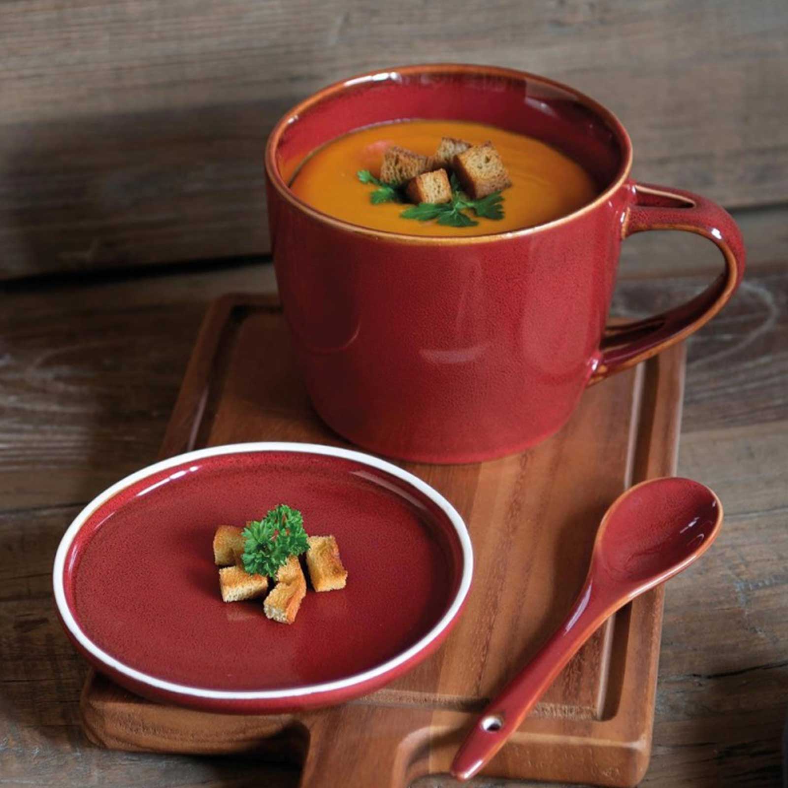 Mug en céramique rouge avec soupe veloutée et croûtons, accompagné d'une petite assiette assortie et d'une cuillère sur un plateau en bois