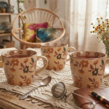 Trois tasses en céramique à motif floral champêtre posées sur un napperon en macramé avec une petite cuillère et un infuseur à thé