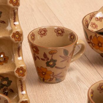 Tasse en grès beige orné de motifs floraux automnaux, posé à côté d'un service à vaisselle assorti dans un style artisanal chaleureux