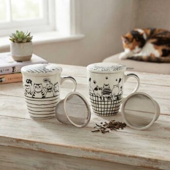 Deux tasses à thé en céramique ornées de motifs de chats avec couvercles et infuseurs métalliques amovibles posées sur une table en bois clair