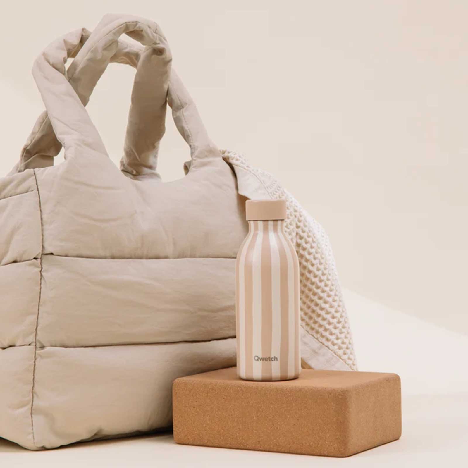 Bouteille isotherme Qwetch beige à rayures blanches posée sur un bloc de yoga, accompagnée d'un sac cabas matelassé dans un environnement minimaliste