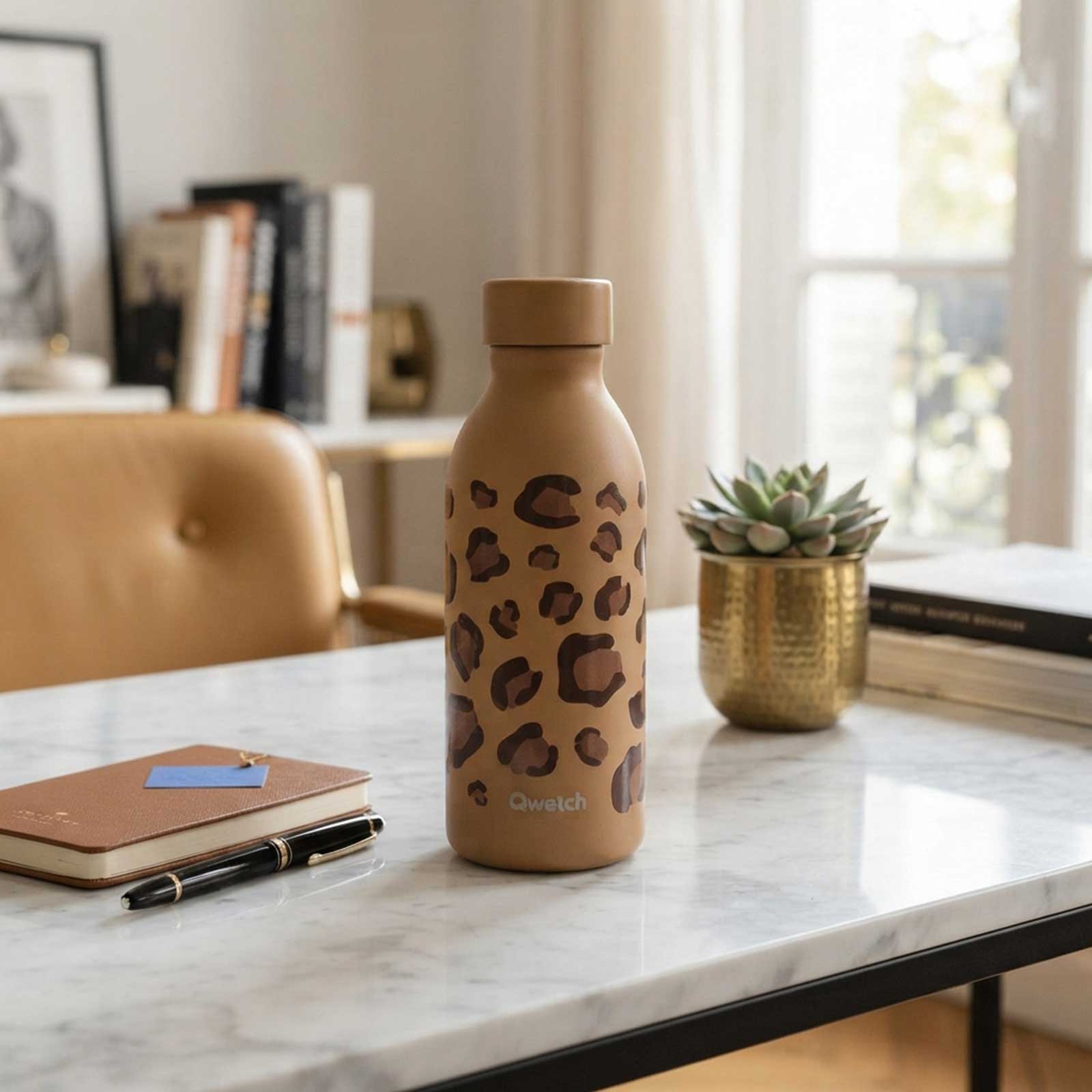 Bouteille isotherme Qwetch à motif léopard posée sur une table en marbre, à côté d'un carnet, d'un stylo et d'une plante succulente en pot doré