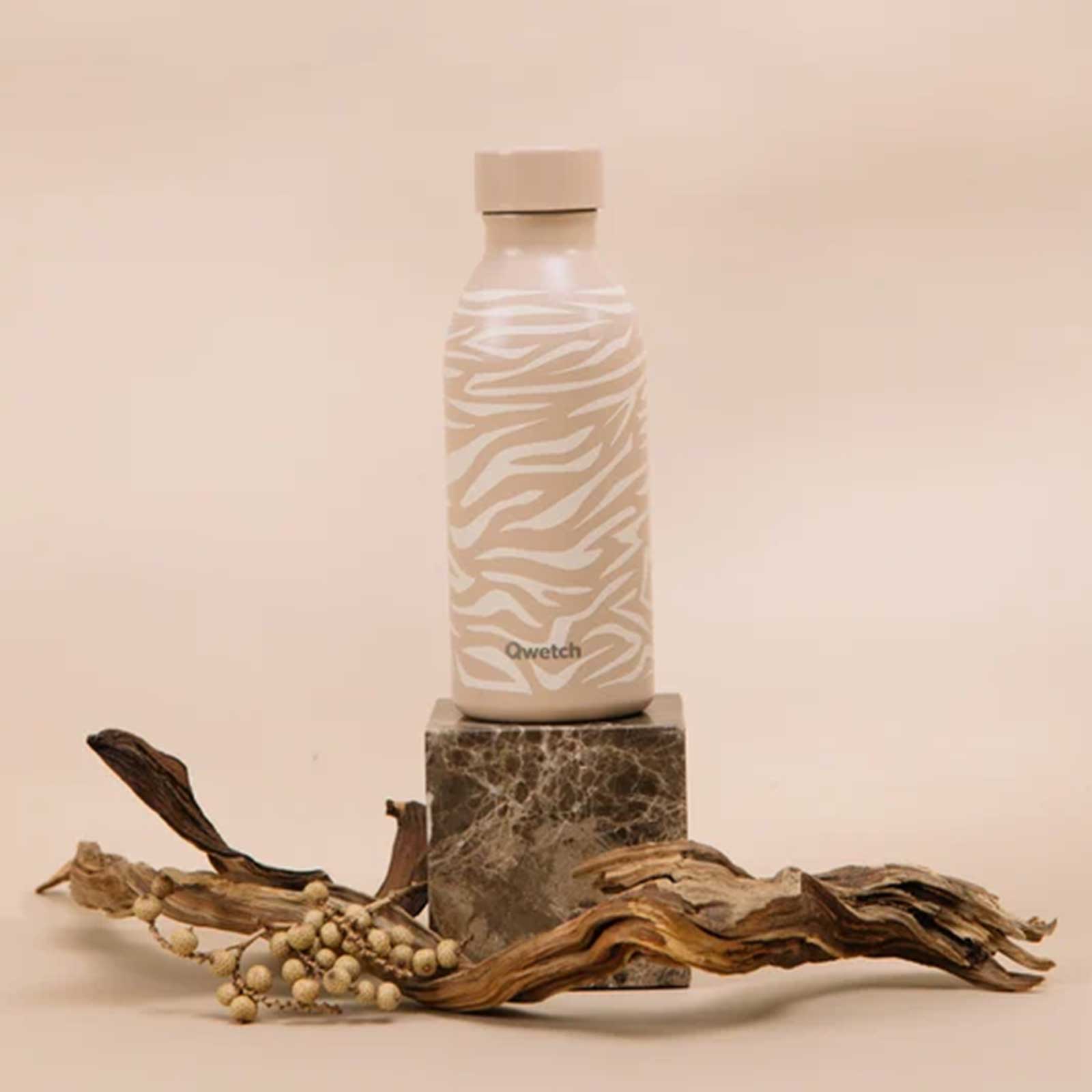 Bouteille isotherme Qwetch avec un motif zébré beige posée sur un cube en marbre au milieu de morceaux de bois flotté et de grappes de fruits secs