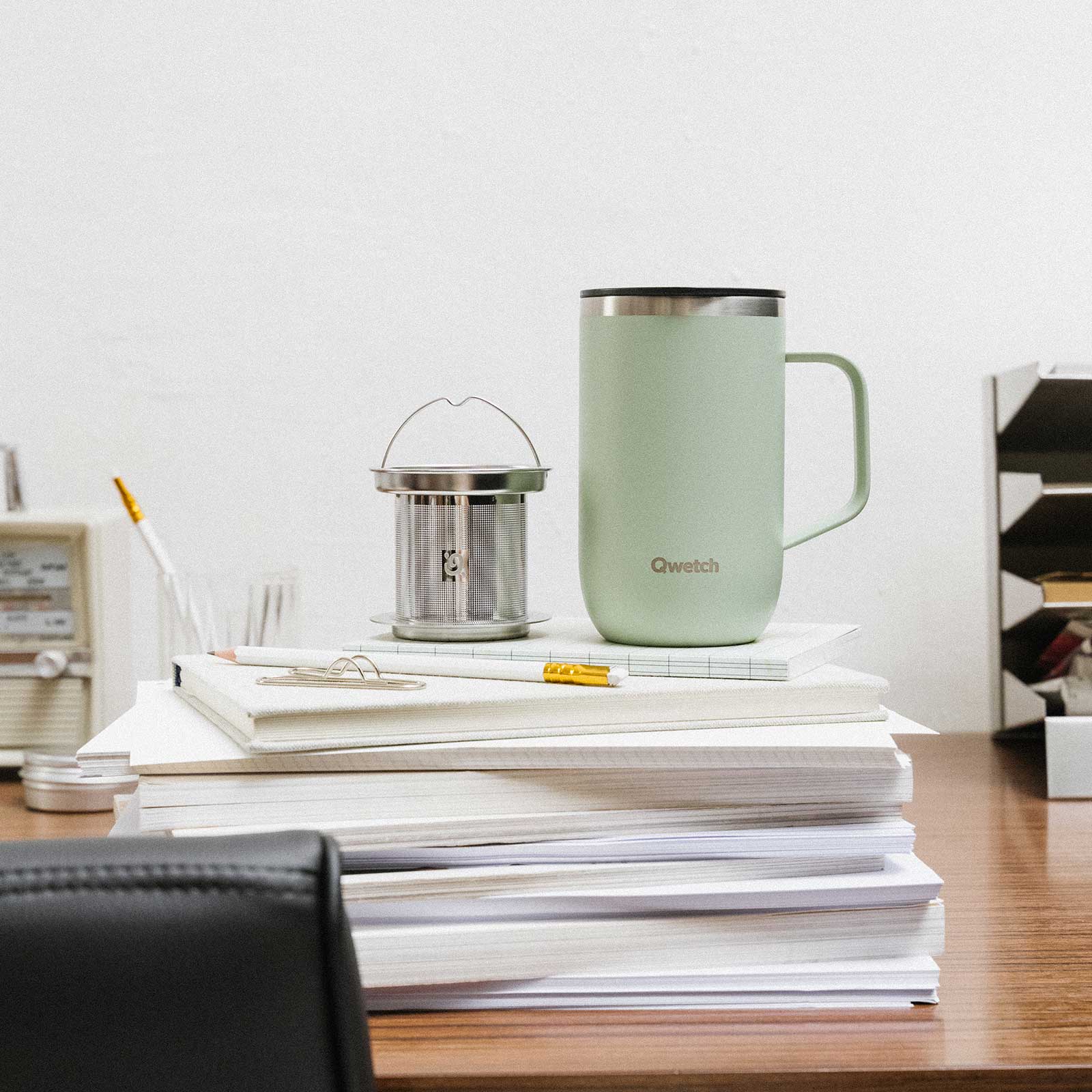 Mug isotherme vert sauge Qwetch avec infuseur en inox posé sur une pile de carnets blancs sur un bureau