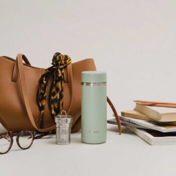 Gourde isotherme vert sauge de marque Etch posée à côté d'un sac à main marron, de lunettes et de livres, accessoires lifestyle pour le quotidien