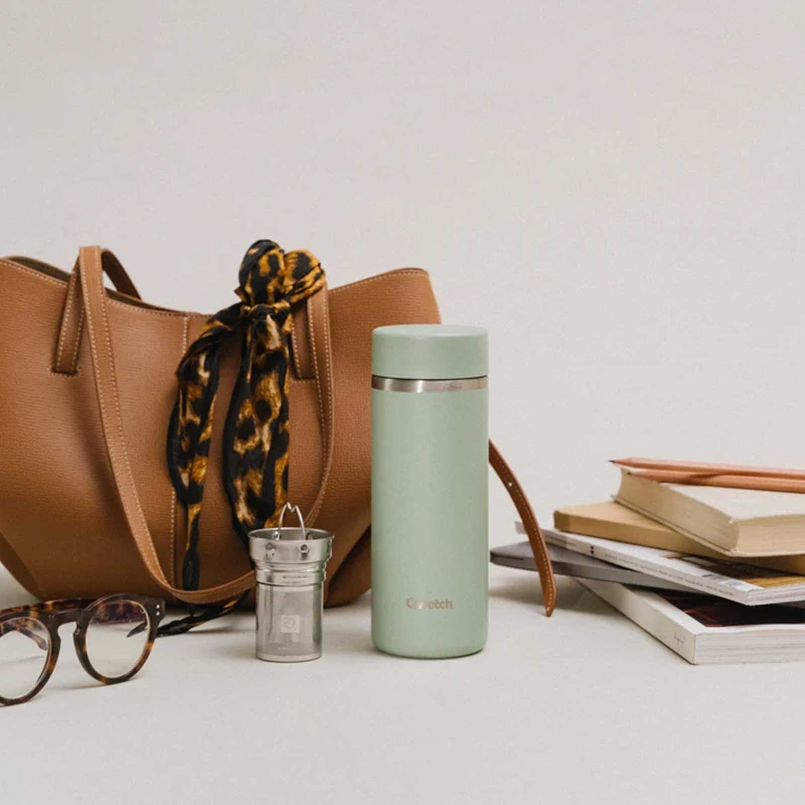 Gourde isotherme vert sauge de marque Etch posée à côté d'un sac à main marron, de lunettes et de livres, accessoires lifestyle pour le quotidien