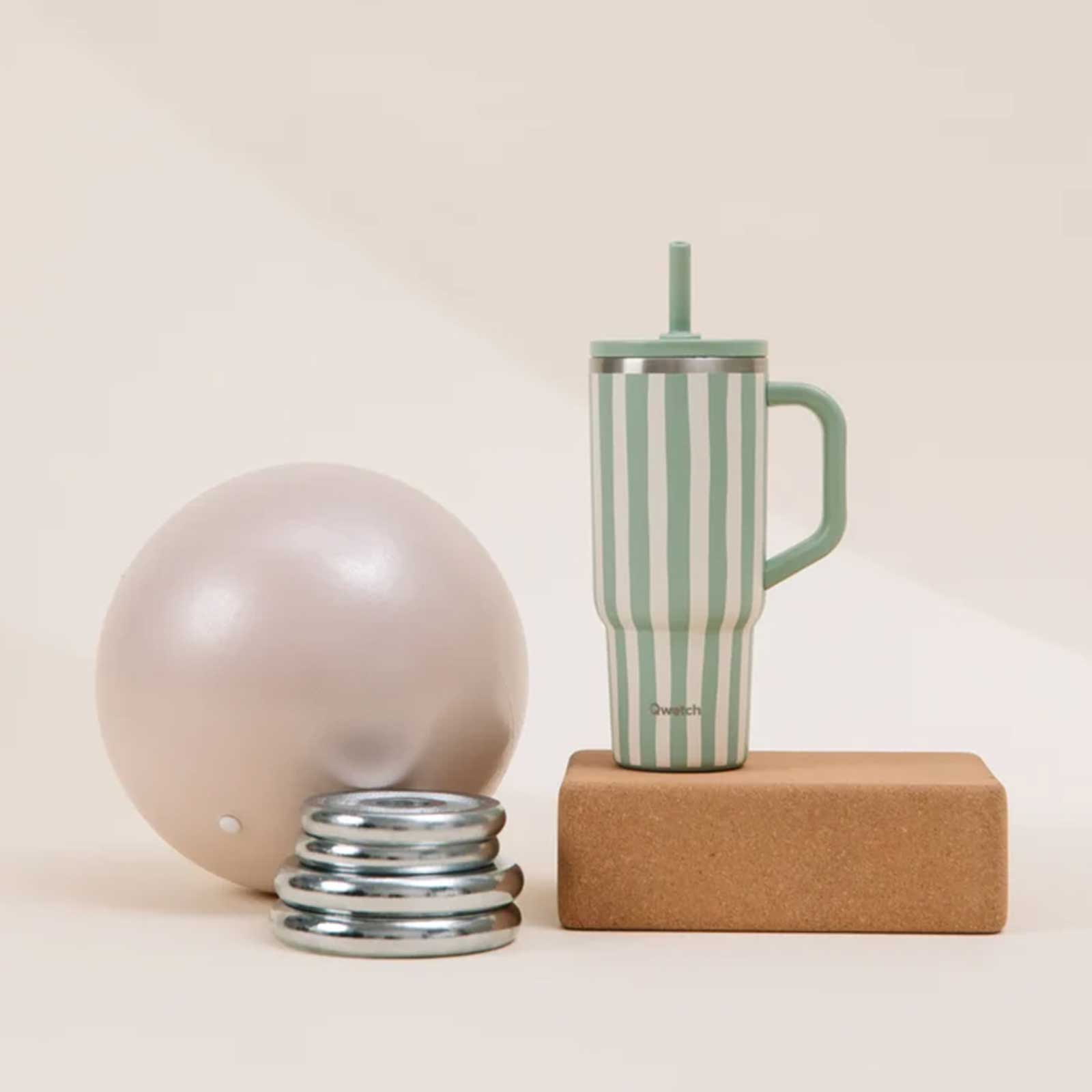 Gourde Qwatch à rayures vertes et blanches posée sur un bloc de yoga en liège, accompagnée d'un ballon et de disques de musculation.