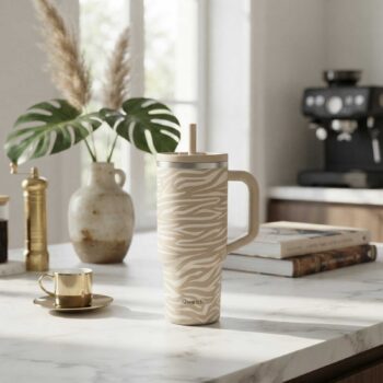 Gobelet isotherme Qwetch beige à motifs zébrés avec paille et anse, posé sur un comptoir en marbre près d'une machine à café