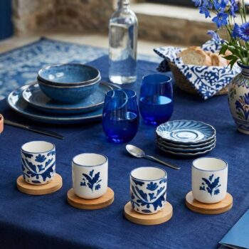 Quatre tasses en céramique peintes de motifs bleus floraux posées sur des sous-verres en bois sur une table dressée avec de la vaisselle bleue