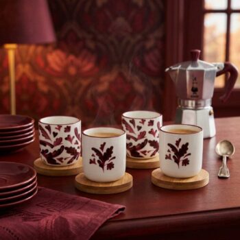 Quatre tasses en céramique décorées de motifs de feuilles automnales posées sur des sous-verres en bois près d'une cafetière italienne