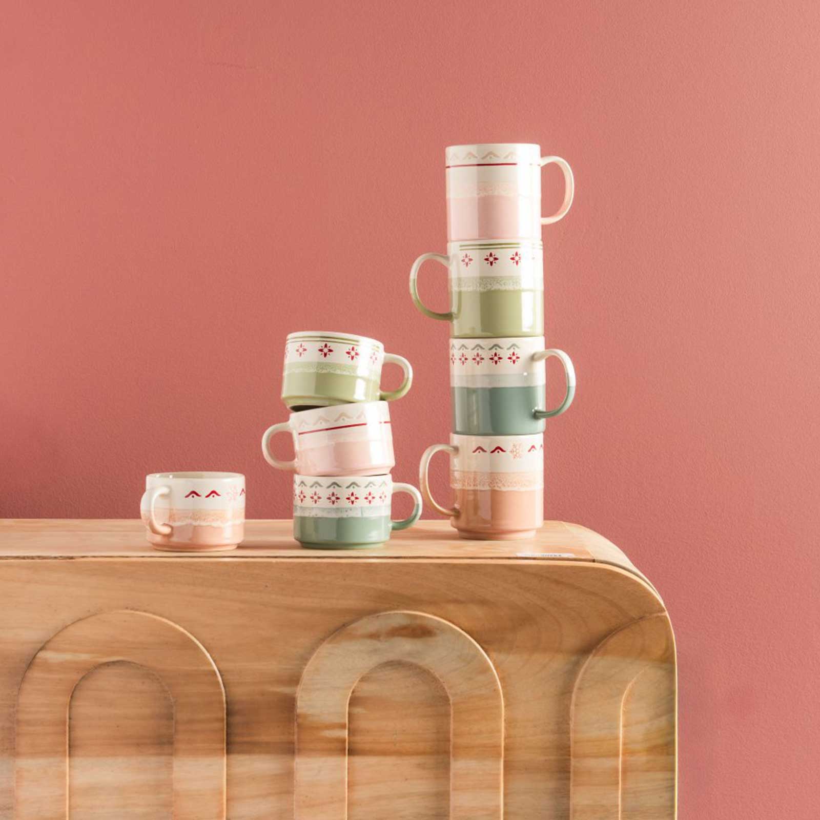 Piles de mugs en céramique aux motifs géométriques et aux couleurs pastel disposées sur une console en bois courbé devant un mur rose