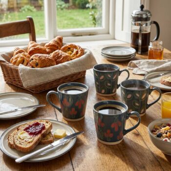 Service de petit-déjeuner sur table en bois avec tasses en céramique à motifs floraux, panier de viennoiseries et tartines à la confiture