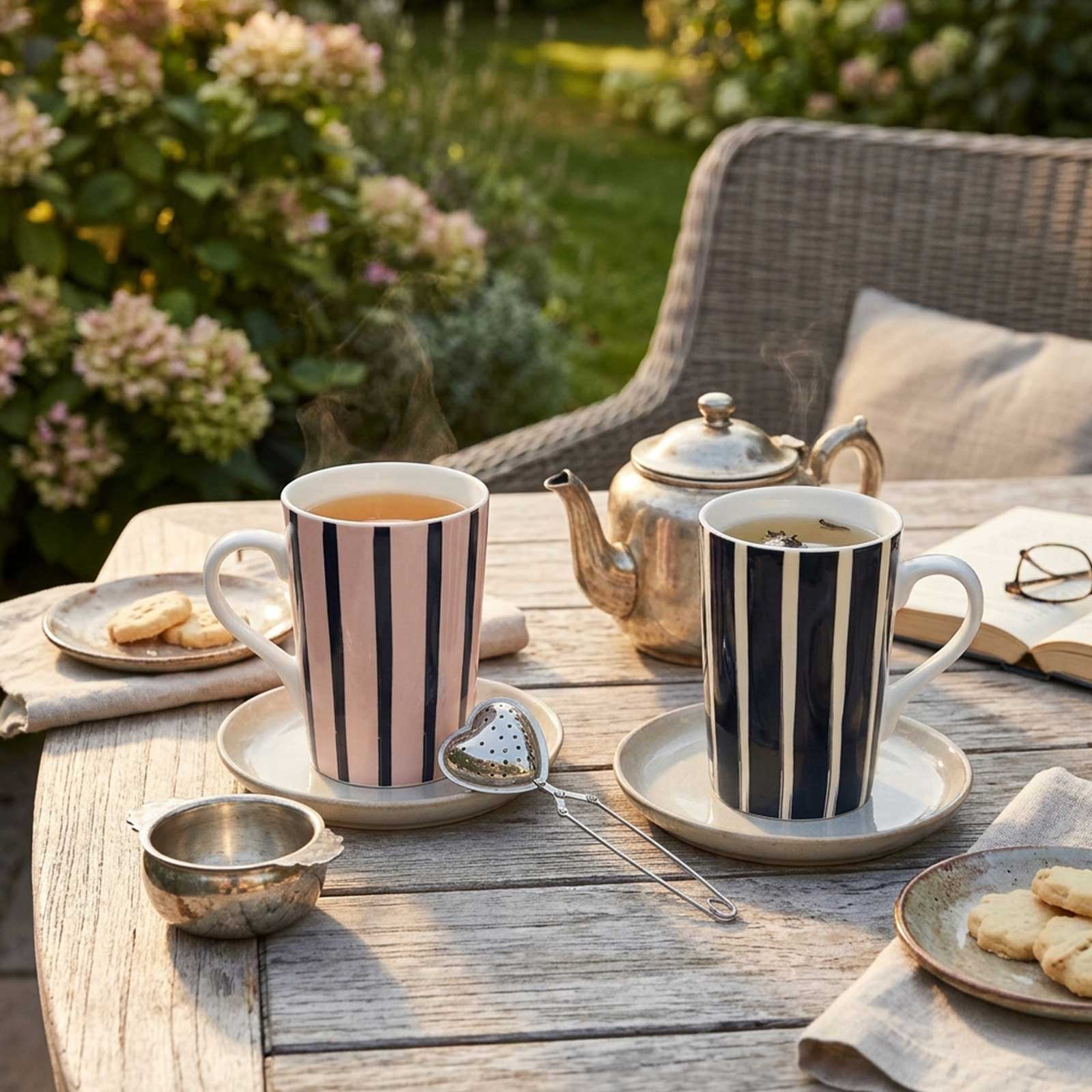 Deux tasses en céramique à rayures verticales posées sur une table en bois avec une théière en argent, une infuseur en cœur et des petits biscuits