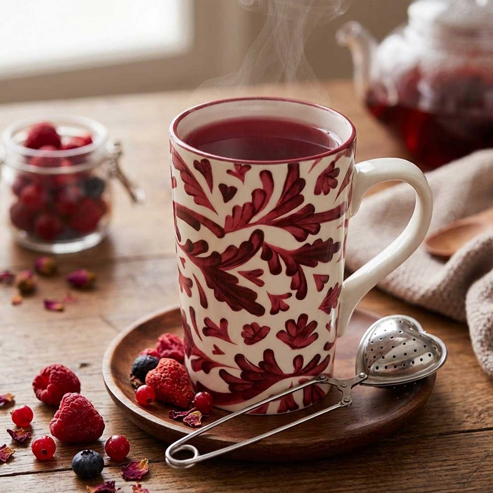 Mug en céramique à motifs végétaux rouges sur une soucoupe en bois, accompagné d'une boule à thé en forme de cœur et de fruits rouges frais