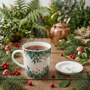 Mug à thé en céramique décoré de branches de sapin et baies rouges posé sur une table en bois avec une théière en cuivre et des champignons décoratifs