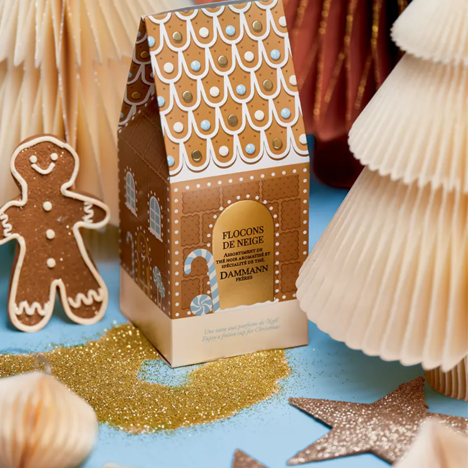 Coffret de thé Dammann Frères en forme de maison en pain d'épices, entouré de sapins en papier et de paillettes dorées pour une ambiance de Noël.