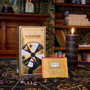 Coffret de thé Kusmi Tea Les Gourmands et sachet de thé Le Dessert posés sur une nappe ésotérique avec une bougie noire et des pierres violettes.