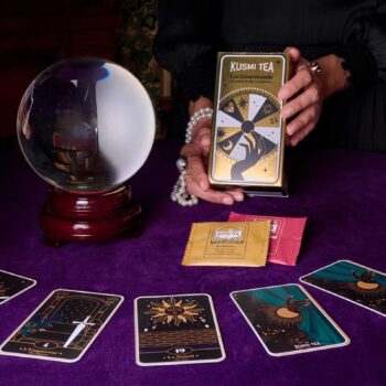Mains tenant une boîte de thé Kusmi Tea Les Gourmands devant une boule de cristal et des cartes de tarot sur une nappe violette