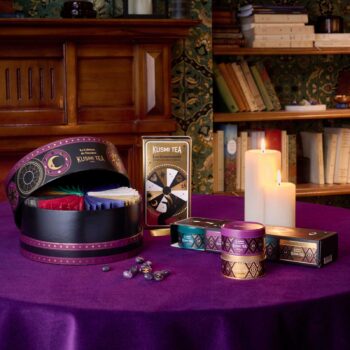 Coffret cadeau Kusmi Tea Le Cabinet de Voyage ouvert sur une table violette, entouré de bougies et de thés assortis devant une bibliothèque.