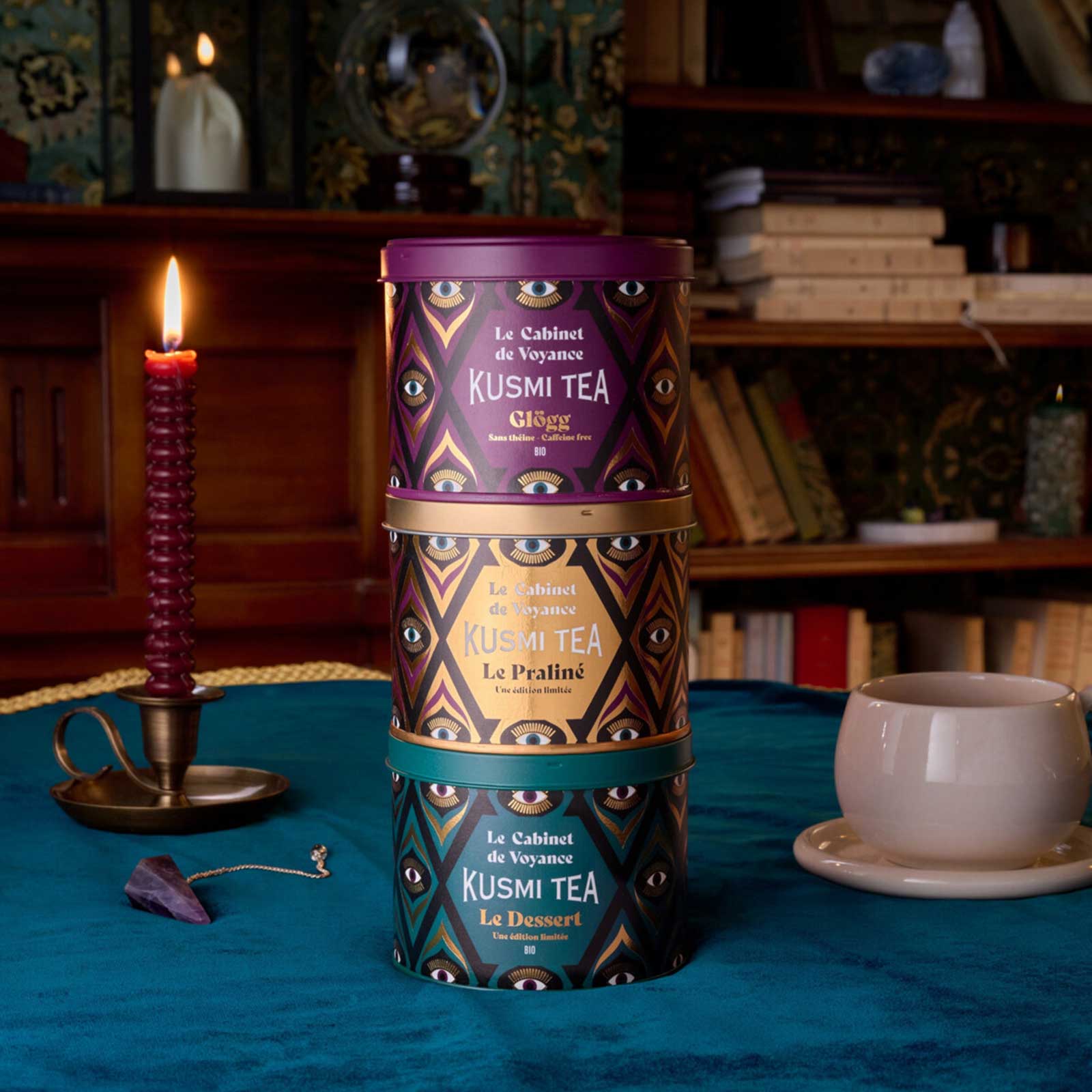 Trois boîtes de thé Kusmi Tea empilées sur une table bleue dans une ambiance feutrée avec une bougie allumée et des livres en arrière-plan
