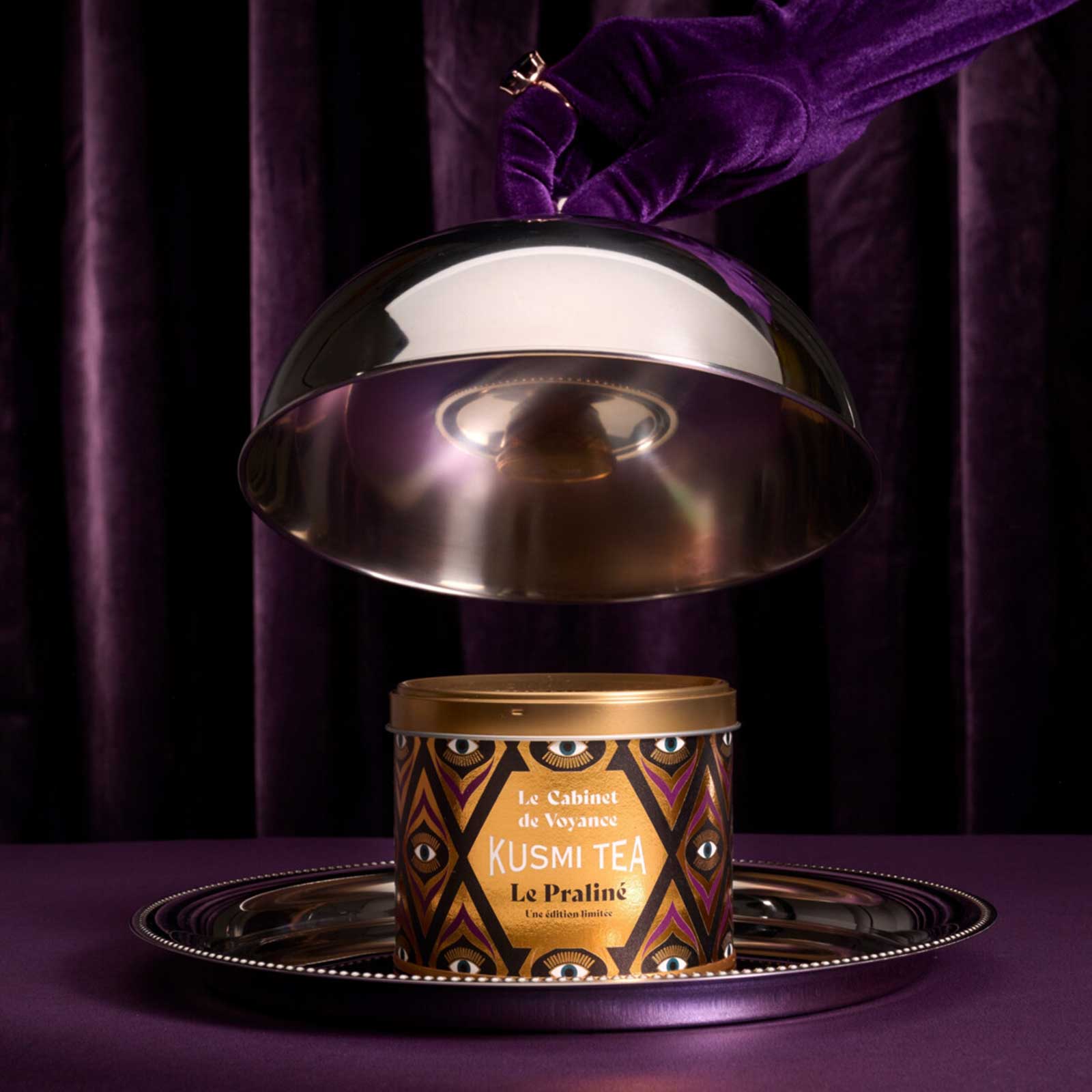 Boîte de thé Kusmi Tea Le Praliné posée sur un plateau argenté sous une cloche soulevée par une main gantée de violet, sur fond de rideaux sombres
