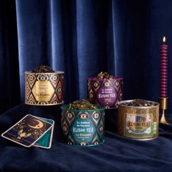 Quatre boîtes de thés Kusmi Tea de la collection Le Cabinet de Voyance disposées devant un rideau bleu sombre avec une bougie et une carte divinatoire
